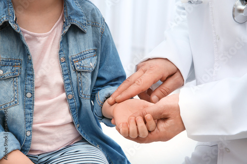 Doctor checking little girl's pulse with fingers on light background, closeup