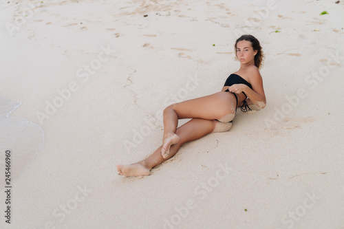 Sexy tanned model in black swimsuit posing on white sandy beach