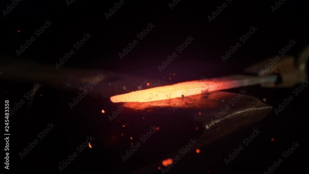 SLOW MOTION, MACRO, DOF: Small black particles flying away from hot red blade while getting forged by an unrecognizable blacksmith. Glowing piece of iron being forged into blade. Forging knife blades.