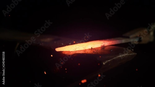 SLOW MOTION, MACRO, DOF: Small black particles flying away from hot red blade while getting forged by an unrecognizable blacksmith. Glowing piece of iron being forged into blade. Forging knife blades.