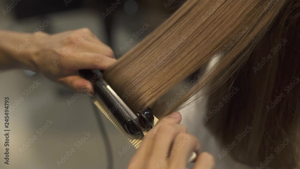 Hairstyling long hair with hair iron in professional beauty studio. Hairstylist doing straightening for long haired woman in barbershop. Hairdresser creating hairstyle brunette woman.