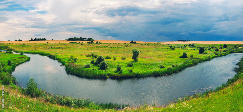 Obraz premium Cloudy summer panoramic landscape with golden fields,green meadows,woods and small river