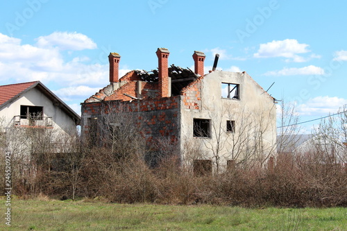 Family house bombarded during war with missing windows and completely destroyed roof with only chimneys left untouched surrounded with overgrown underbrush and cloudy blue sky background