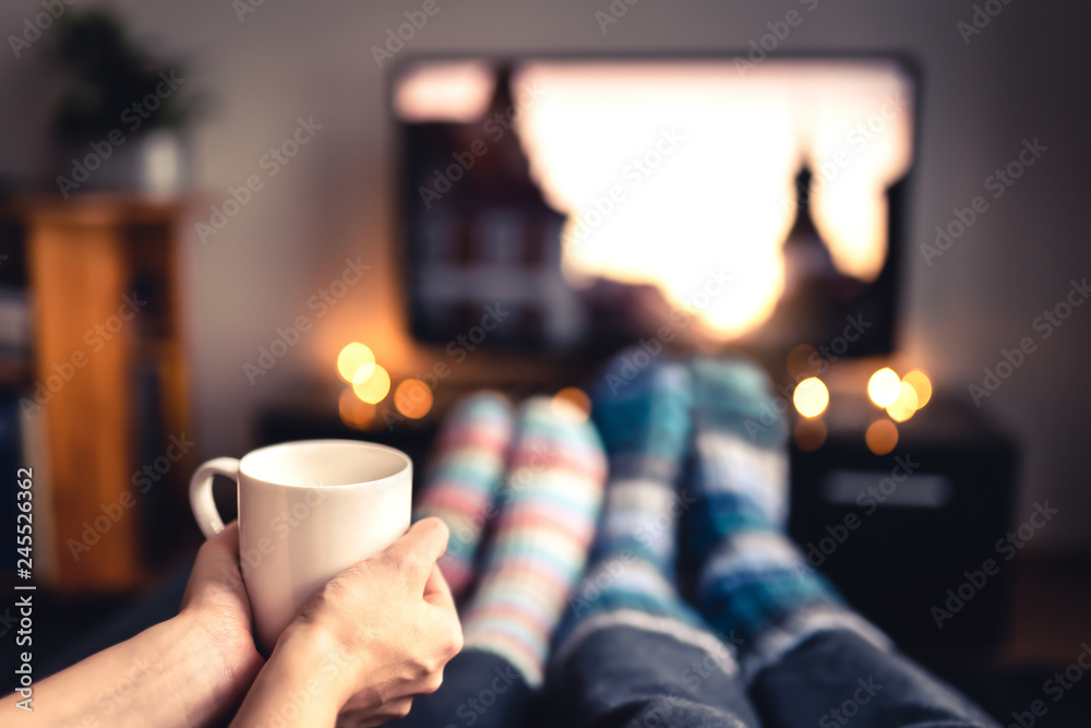 Couple drinking tea, hot chocolate, eggnog or mulled wine and watching tv in warm cozy woolen socks in winter. Woman holding cup of morning coffee in home living room. Sick people with flu or fever. - obrazy, fototapety, plakaty 