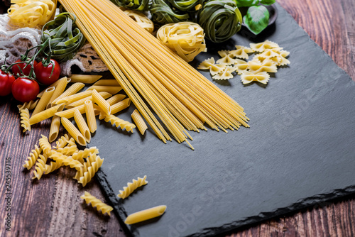 Assortment of many different raw pasta on a rustic board, beautifully arranged with basil and tomatoes