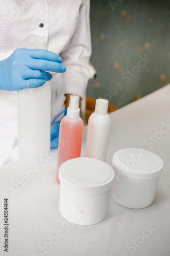 Close up of female hands in latex gloves with cosmetic and personal care products on table. Lady is wearing white lab coat. concept of professional beauty products