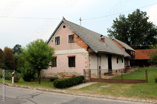 Small old suburban family house with broken facade and dilapidated windows surrounded with freshly cut grass and trees with paved road in front