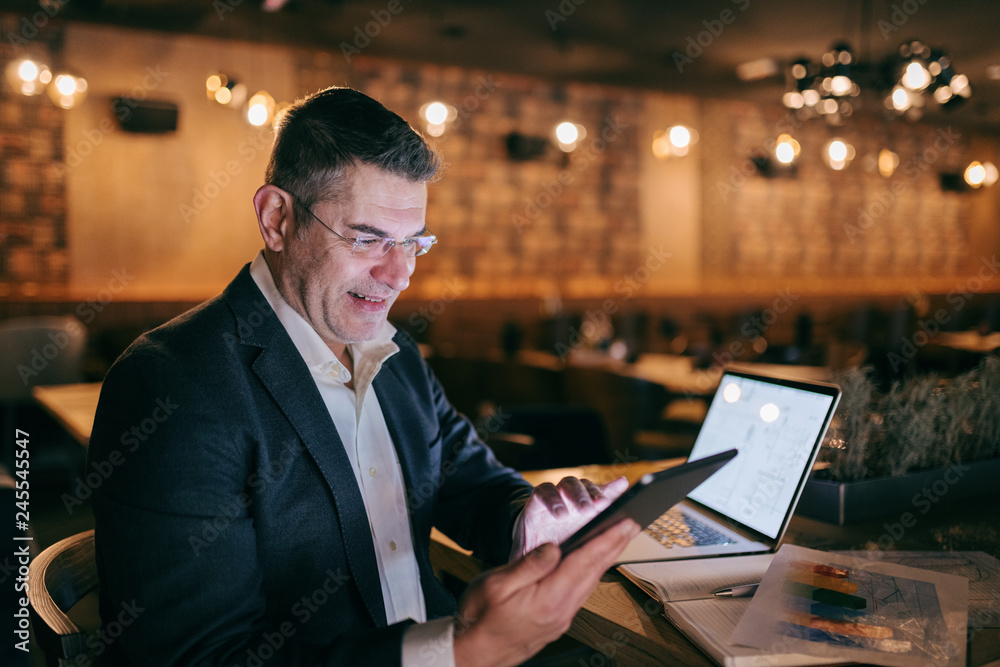 Middle-aged businessman dressed smart casual using tablet while siting in cafe. On the desk laptop and paperwork. Middle-aged businessman dressed smart casual using tablet while siting in cafe. On the desk laptop and paperwork.