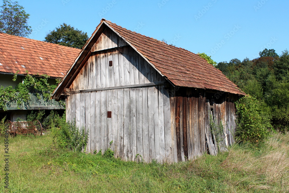 Unused old abandoned dilapidated wooden barn with boarded entrance and cracked boards surrounded with high uncut grass and trees with rusted agricultural equipment and old house in background