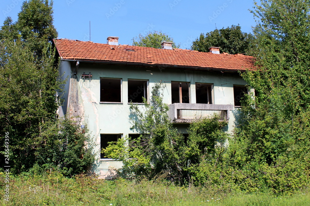 War destroyed abandoned building with dilapidated facade and missing doors and windows completely surrounded with overgrown high grass and forest vegetation with clear blue sky in background