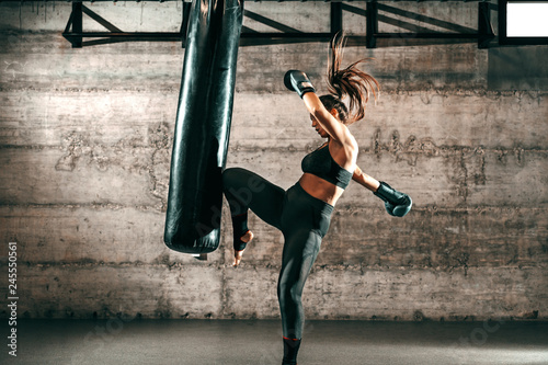 Fotografie, Obraz Dedicated strong brunette with ponytail, in sportswear, bare foot and with boxing gloves kicking sack in gym