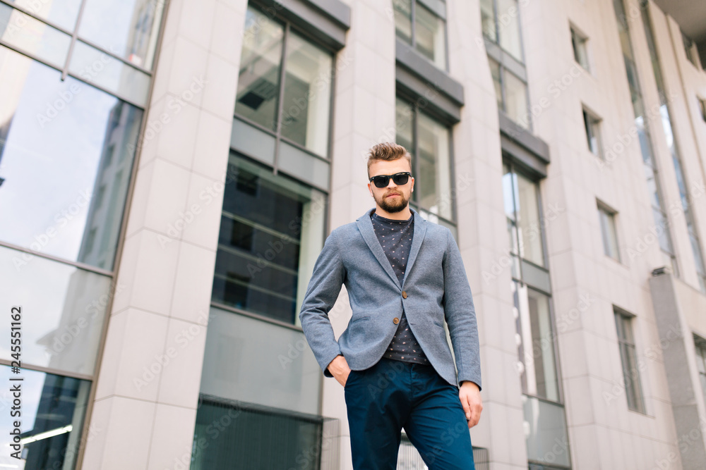 Handsome guy with beard is standing outdoor on office building background. He wears gray jacket, jeans, sunglasses. He looks to the camera. Bottom view. Handsome guy with beard is standing outdoor on office building background. He wears gray jacket, jeans, sunglasses. He looks to the camera. Bottom view.