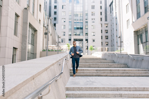 Full-length photo of  man in sunglasses walking on concrete stairs on  office building background. He wears T-shirt, jacket,  jeans, brown shoes. © Look!