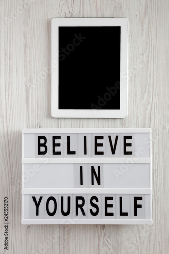 Modern board with text 'Believe in yourself', tablet on a white wooden background, top view. From above, flat lay, overhead.