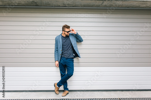 Full-length photo handsome guy posing on gray wall background. He wears gray jacket, T-shirt, jeans, brown shoes. He is holding sunglasses on face and smiling to the side.