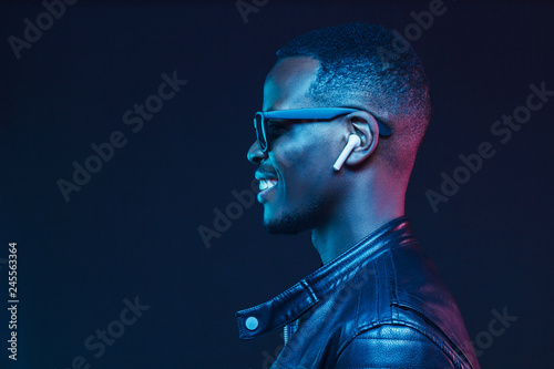 Neon portrait of smiling african man listening music with earphones, wearing black leather jacket