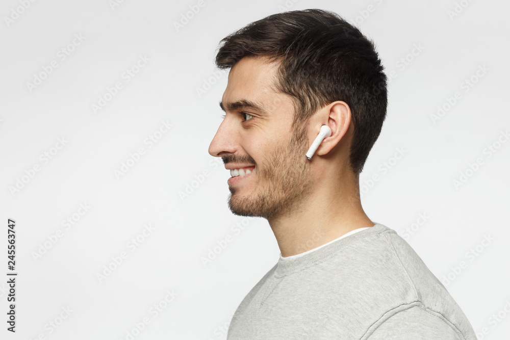 Fototapeta premium Sideways portrait of smiling young man listening to music or radio, uses modern wireless earphones