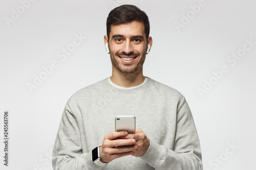 Young man standing isolated on gray background, looking at camera with smile, holding smartphone with both hands