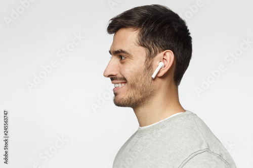 Sideways portrait of smiling young man listening to music or radio, uses modern wireless earphones