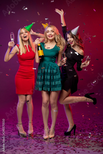 Portrait of three amazing, cute girls in fashion dresses and party caps, holding glass with champagne, celebrating the New year or Birthday party, standing over colorful background