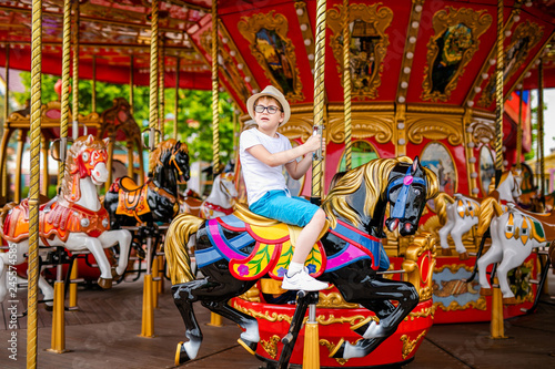 Blonde boy in the straw hat and big glasses riding colorful horse in the merry-go-round carousel in the entertainment park © Lena May