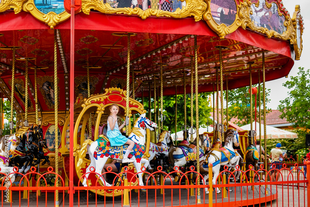 Blonde girl with two braids in white and blue dress riding colorful horse in the merry-go-round carousel in the entertainment park Blonde girl with two braids in white and blue dress riding colorful horse in the merry-go-round carousel in the entertainment park