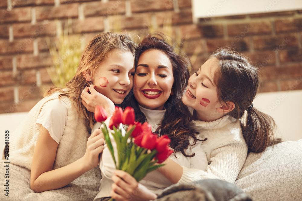 Amazed cheerful joyful charming mum is holding nice flowers and is giving kisses to her little cute daughters who is congratulating mother's day to her. Amazed cheerful joyful charming mum is holding nice flowers and is giving kisses to her little cute daughters who is congratulating mother's day to her.
