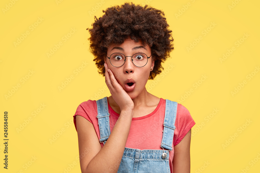 Stupefied young dark skinned curly lady shocked with mess on workplace, keeps jaw dropped, touches cheek with palm, wears pink t shirt and denim overalls, isolated over yellow studio background - obrazy, fototapety, plakaty 