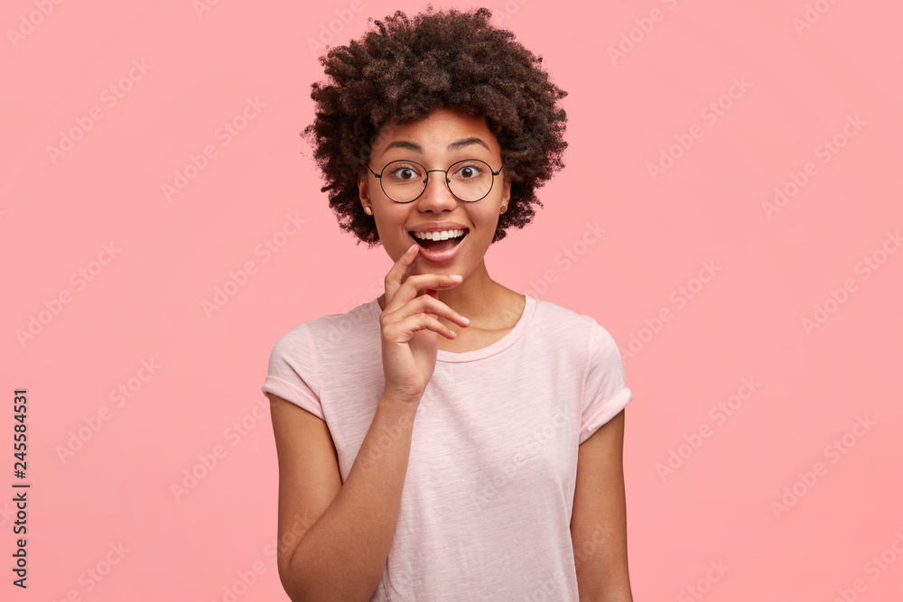 Headshot of attractive cheerful woman with curly hair, wears transparent glasses, smiles happily, dressed in casual t shirt, stands against pink background, feels optimistic, hears praise. Emotions Headshot of attractive cheerful woman with curly hair, wears transparent glasses, smiles happily, dressed in casual t shirt, stands against pink background, feels optimistic, hears praise. Emotions
