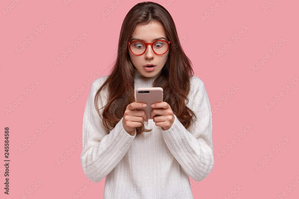 Photo of fearful dark haired lady stares at cell phone with surprised expression wears casual white sweater, reads shocking news on internet website, isolated over pink background. Astonishment Photo of fearful dark haired lady stares at cell phone with surprised expression wears casual white sweater, reads shocking news on internet website, isolated over pink background. Astonishment
