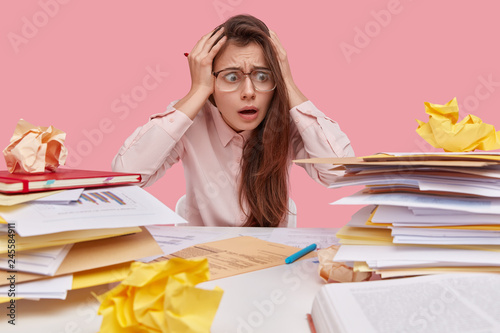 Worried overwhelmed young woman stares at pile of papers, keeps hands on head, feels nervous before business meeting, prepares financial report, isolated over pink background, poses at workplace photo