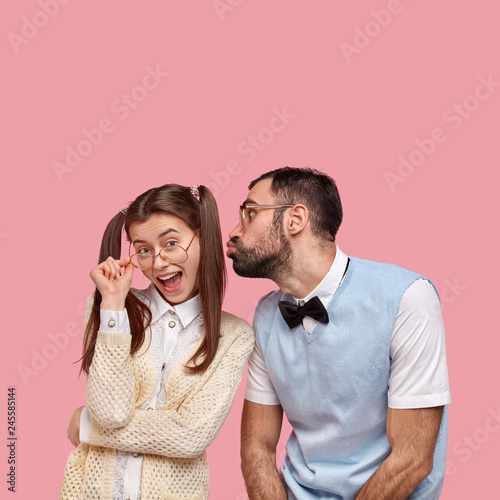 Joyful woman dressed in old fashionable clothes, keeps hand on frame of glasses, flirts with someone, looks happily through spectacles, clumsy man with bristle, wears vest and bowtie, wants to kiss photo