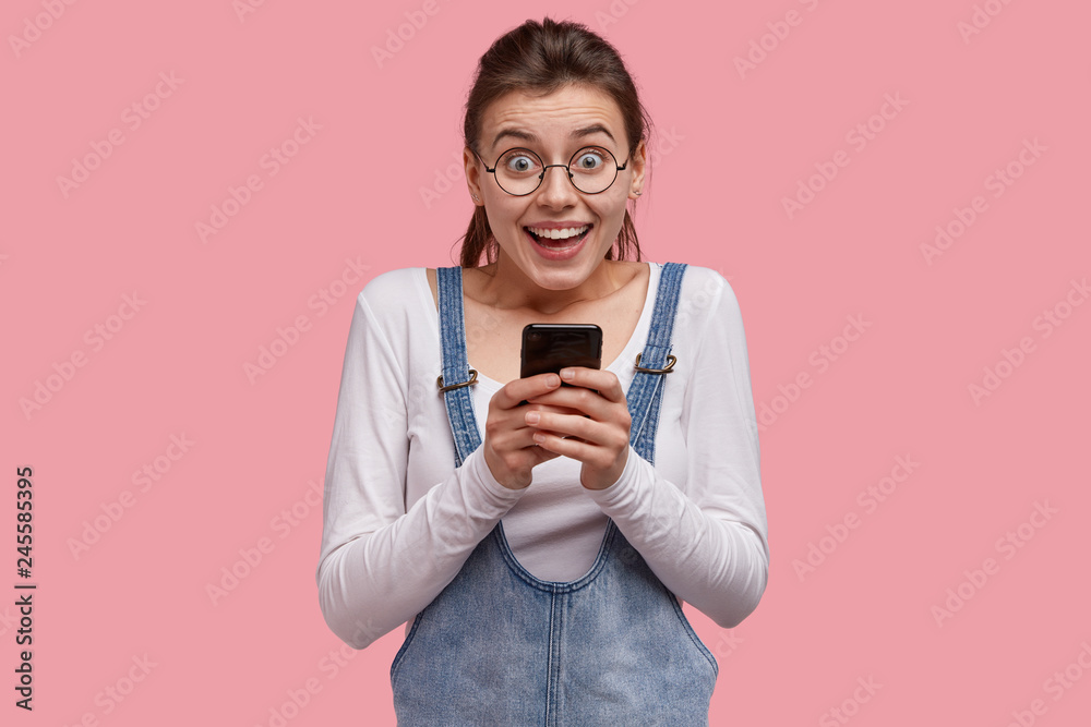 Modern technologies and communication concept. Joyful dark haired lovely woman in round spectacles, holds cell phone, has positie expression, wears casual outfit, sends text message to boyfriend Modern technologies and communication concept. Joyful dark haired lovely woman in round spectacles, holds cell phone, has positie expression, wears casual outfit, sends text message to boyfriend