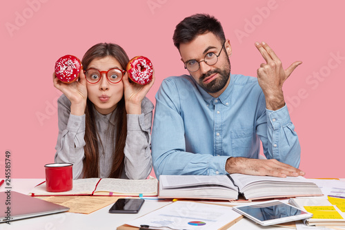 Upset overworked bearded man shoots in temple, makes suicide gesture, wears spectacles and elegant shirt, attractive lady in eyewear holds red donuts near head, read and write information together