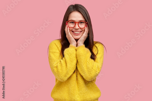 Happy woman keeps hands on cheeks, smiles amiably, has fun chatting with friends, wears spectacles and yellow clothes, isolated over pink background, talks to boy she likes. Good mood concept photo