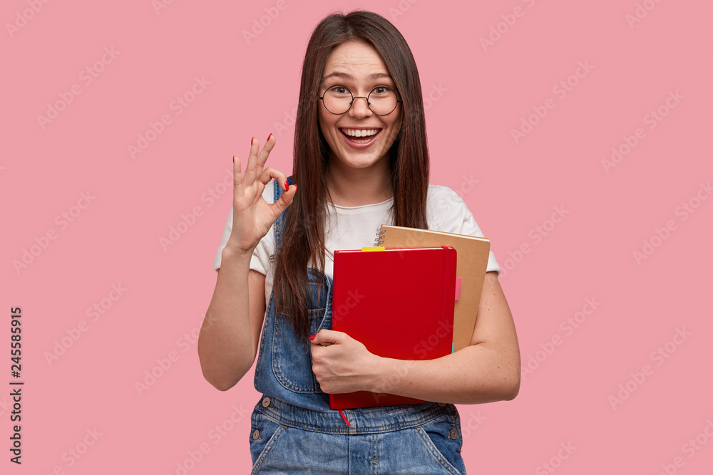 Happy brunette woman agrees with list to do, carries necessary notepad for writing, makes okay gesture, dressed in denim overalls and t shirt, isolated over pink background. Agrreement and approval Happy brunette woman agrees with list to do, carries necessary notepad for writing, makes okay gesture, dressed in denim overalls and t shirt, isolated over pink background. Agrreement and approval