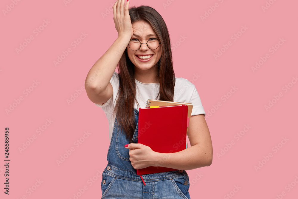 Pleased college student keeps hand on head, smiles broadly, holds textbook, remember about important task, being in good mood, wears denim dungarees and white t shirt, models indoor. Studying concept Pleased college student keeps hand on head, smiles broadly, holds textbook, remember about important task, being in good mood, wears denim dungarees and white t shirt, models indoor. Studying concept