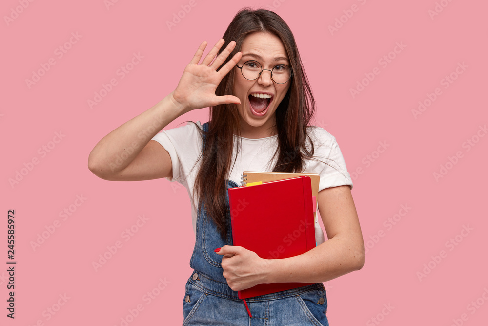 Joyful brunette woman waves with palm, wears optical glasses and denim overalls, exclaims in happiness, holds spiral notepad, models against pink background. Happy student greets with friend Joyful brunette woman waves with palm, wears optical glasses and denim overalls, exclaims in happiness, holds spiral notepad, models against pink background. Happy student greets with friend