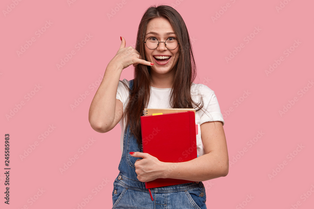 Positive young woman with cheerful expression, makes call gesture being in high spirit smiles broadly carries organizer poses against pink background. Teenage girl asks friend to call back on distance Positive young woman with cheerful expression, makes call gesture being in high spirit smiles broadly carries organizer poses against pink background. Teenage girl asks friend to call back on distance