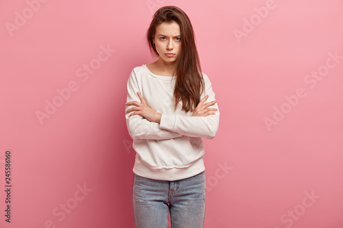 Irritated young exasperate woman keeps arms crossed, strongly dissatisfied with situation, has frowned eyebrows, demonstrates anger, wears casual jumper and jeans, stands against pink background © wayhome.studio 