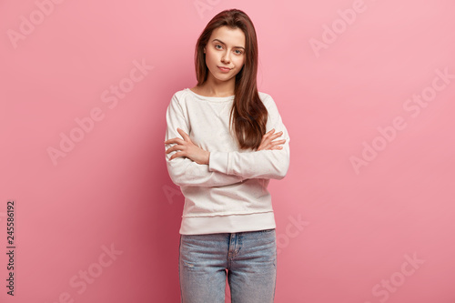 Image of pleasant looking self assured woman with serious expression, keeps arms folded, listens information attentively, dressed in sweatshirt, jeans, isolated over pink background, being thoughtful