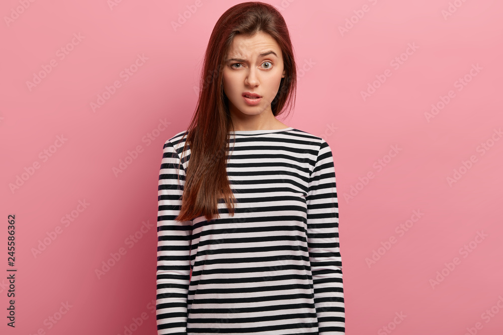Indignant dissatisfied woman raises eyebrows, has surprised expression, wears striped clothes, expresses dislike, has puzzled reaction, isolated over pink background. Human facial expressions concept Indignant dissatisfied woman raises eyebrows, has surprised expression, wears striped clothes, expresses dislike, has puzzled reaction, isolated over pink background. Human facial expressions concept