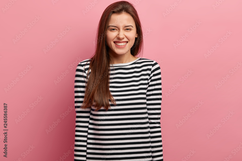 Fototapeta premium Pleased pleasant looking woman giggles as sees something awesome, has dark straight long hair, wears striped outfit, models against pink background, feels carefree, listens joke or funny anecdote