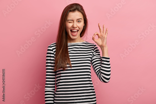 Photo of cheerful young woman blinks eye, makes okay gesture, demonstrates her agreement, feels happy, wears black and white striped jumper, isolated over pink background. Body language concept photo