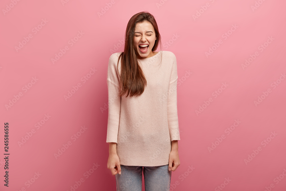 Emotions and feelings concept. Overjoyed pleasant looking young woman laughs loudly, has fun in company of friends, keeps eyes closed with happiness, wears long loose sweater, models indoor. Emotions and feelings concept. Overjoyed pleasant looking young woman laughs loudly, has fun in company of friends, keeps eyes closed with happiness, wears long loose sweater, models indoor.