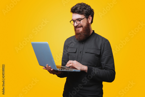 Portrait of cheerful beaded man in glasses, using his laptop over yellow background