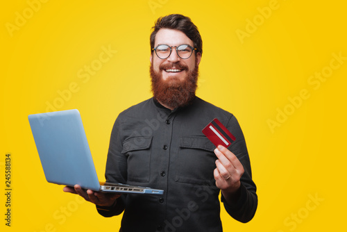 Happysmiling bearded man looking at camera while holding a laptop and showing his credit card