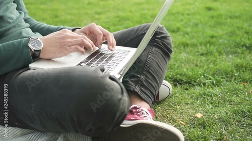 Close up hands of african college student surfing internet sitting on grass. Young man using laptop computer for browsing, communication and studying.