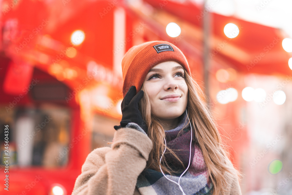 Smiling young woman in warm clothes and a smartphone in her hands listens to music in the headphones and looks sideways on the background of a tourist red bus. Tourist girl walking around the city.