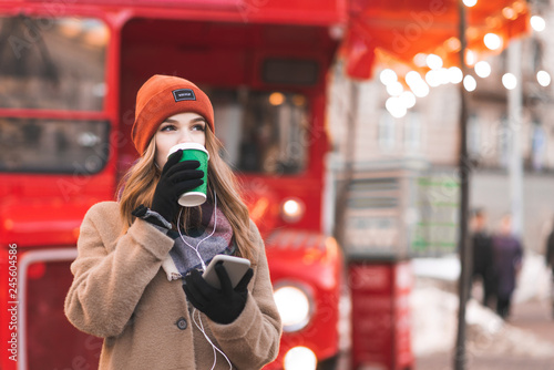 Street portrait of a young woman in warm clothes, standing on the background of a red bus with a smartphone in her hands, drinking coffee from a green cup and looking sideways. Copyspace